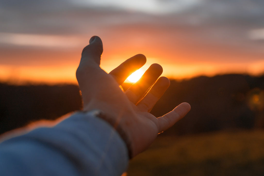 Hand reaching out toward sun setting.