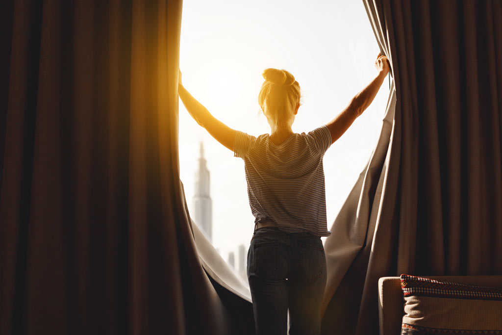 Woman pulling back curtains to allow sunshine in room.